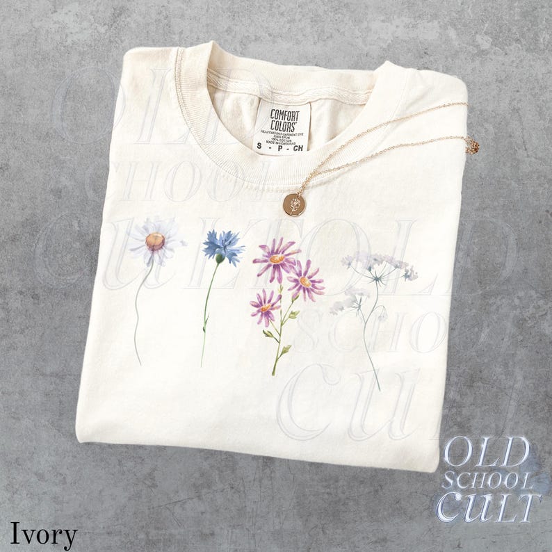 Pu&ograve; includere: T-shirt color avorio con un design floreale ad acquerello con margherite, fiordalisi e altri fiori di campo. Una collana d'oro con un ciondolo &egrave; appoggiata sulla maglietta. La maglietta ha un'etichetta Comfort Colors.