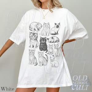 May include: White t-shirt featuring a collection of illustrated cats in various poses. The graphic includes a variety of cat breeds and sizes, all in a black and white sketch style. The text "OLD SCHOOL CULT" is printed on the bottom right.