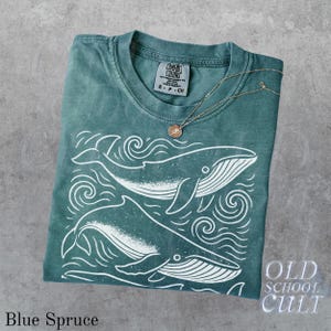 May include: Blue Spruce t-shirt with a whale graphic. The shirt features a crew neck and short sleeves. A gold-colored necklace with a pendant rests on the shirt. The shirt has the text "Comfort Colors" and "Old School Cult".