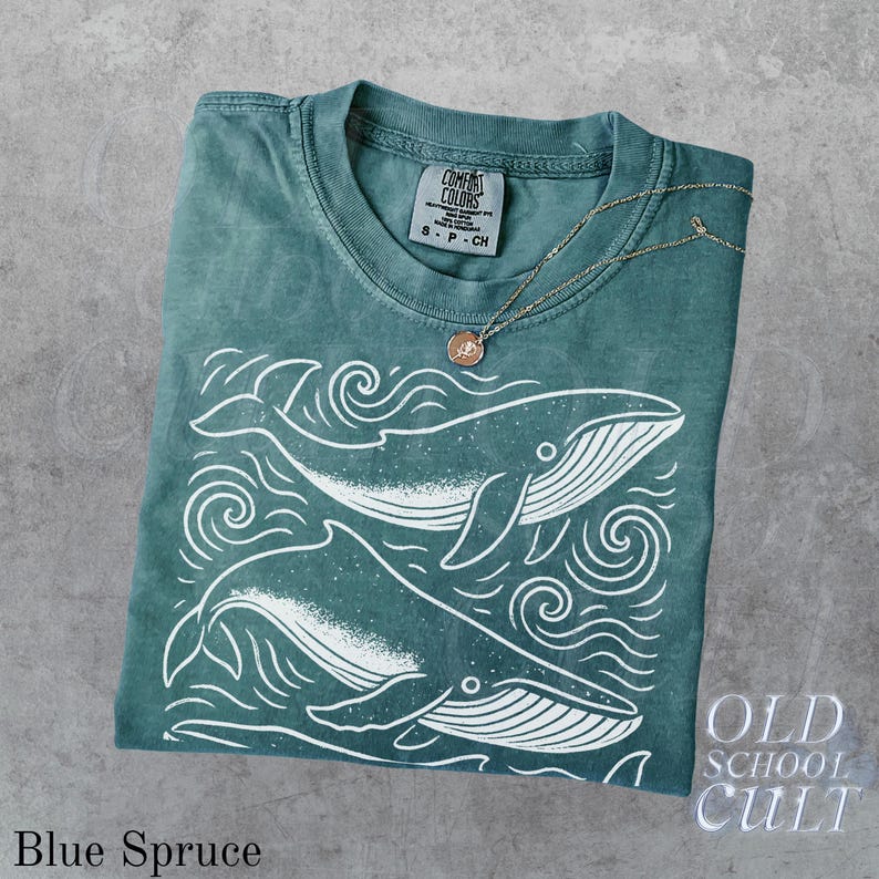 May include: Blue Spruce t-shirt with a whale graphic. The shirt features a crew neck and short sleeves. A gold-colored necklace with a pendant rests on the shirt. The shirt has the text "Comfort Colors" and "Old School Cult".