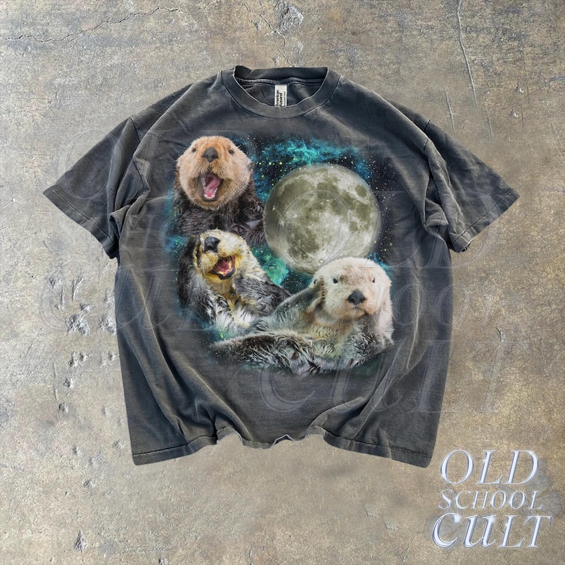 May include: A dark gray t-shirt with a graphic print of three sea otters swimming in front of a full moon. The otters are smiling and appear to be enjoying the night sky.