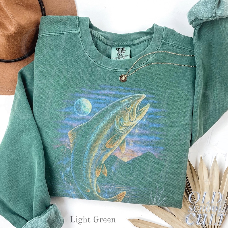 May include: Light green sweatshirt with a graphic of a leaping fish, moon, and mountain scene. The crew neck sweatshirt has long sleeves. A gold necklace with a small pendant is draped on the shirt. The text "Light Green" is at the bottom.