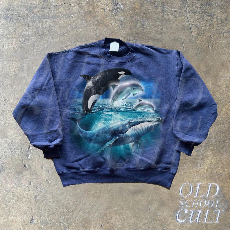 May include: A navy blue sweatshirt with a graphic print of a whale and dolphins jumping out of the water. The graphic is in shades of blue and white.