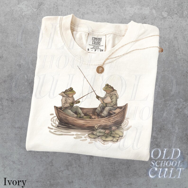 Pode incluir: Camiseta de cor marfim com uma ilustra&ccedil;&atilde;o fantasiosa de dois sapos pescando em um barco de madeira. Os sapos est&atilde;o usando moletons com capuz e s&atilde;o cercados por nen&uacute;fares. Um colar de ouro com um pingente repousa sobre a camiseta. A camiseta tem a etiqueta Comfort Colors.