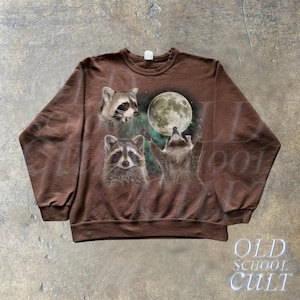 Op de afbeelding: Een bruin sweatshirt met een grafische print van drie wasberen die naar een volle maan kijken. De wasberen zijn omringd door een groene en blauwe nevelachtige achtergrond.