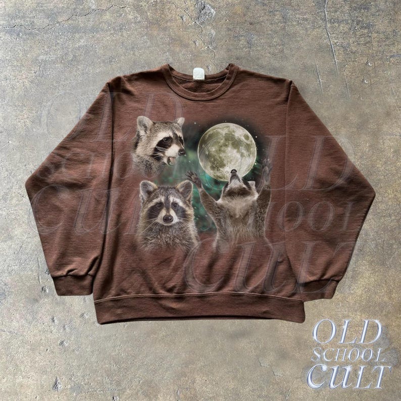Op de afbeelding: Een bruin sweatshirt met een grafische print van drie wasberen die naar een volle maan kijken. De wasberen zijn omringd door een groene en blauwe nevelachtige achtergrond.