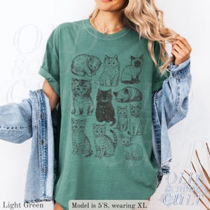 May include: Light green t-shirt featuring a grid of illustrated cats in various poses. The shirt has rolled-up sleeves and is paired with a denim jacket. The text "OLD SCHOOL CULT" is visible on the bottom right.
