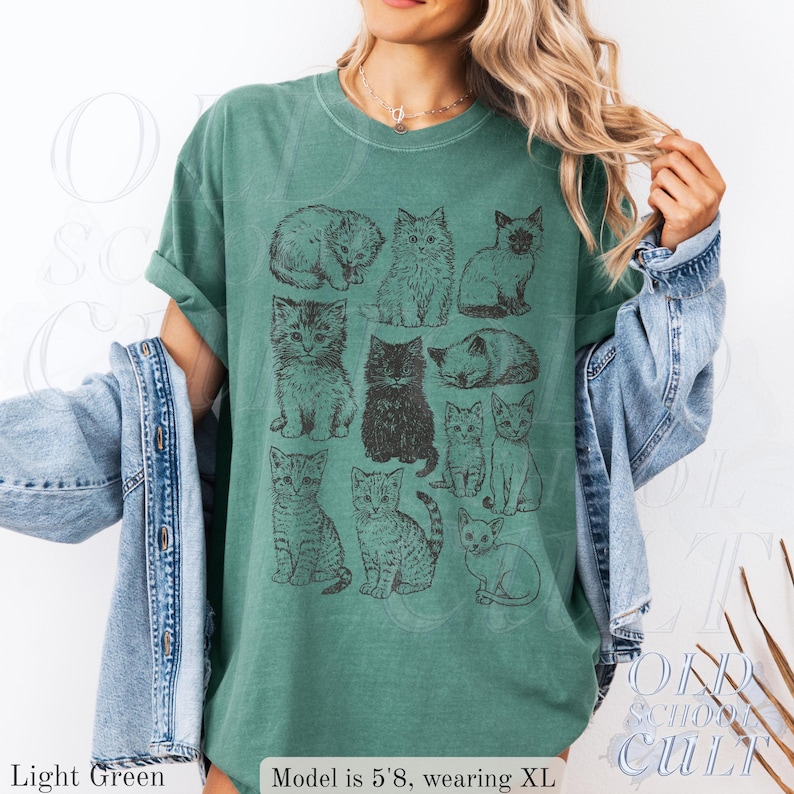 May include: Light green t-shirt featuring a grid of illustrated cats in various poses. The shirt has rolled-up sleeves and is paired with a denim jacket. The text "OLD SCHOOL CULT" is visible on the bottom right.