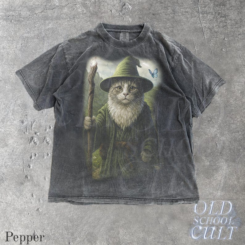 May include: Dark gray t-shirt featuring a cat dressed as a wizard, holding a staff. The cat wears a green hat and robe, with a butterfly nearby. The shirt has the text "OLD SCHOOL CULT" and "Pepper".