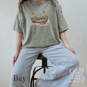 Pode incluir: Camiseta verde s&aacute;lvia com uma ilustra&ccedil;&atilde;o fantasiosa de dois sapos pescando em um barco de madeira. O gr&aacute;fico inclui nen&uacute;fares e varas de pesca. A camiseta &eacute; combinada com cal&ccedil;as de moletom cinza. A palavra "Bay" est&aacute; impressa na parte inferior.