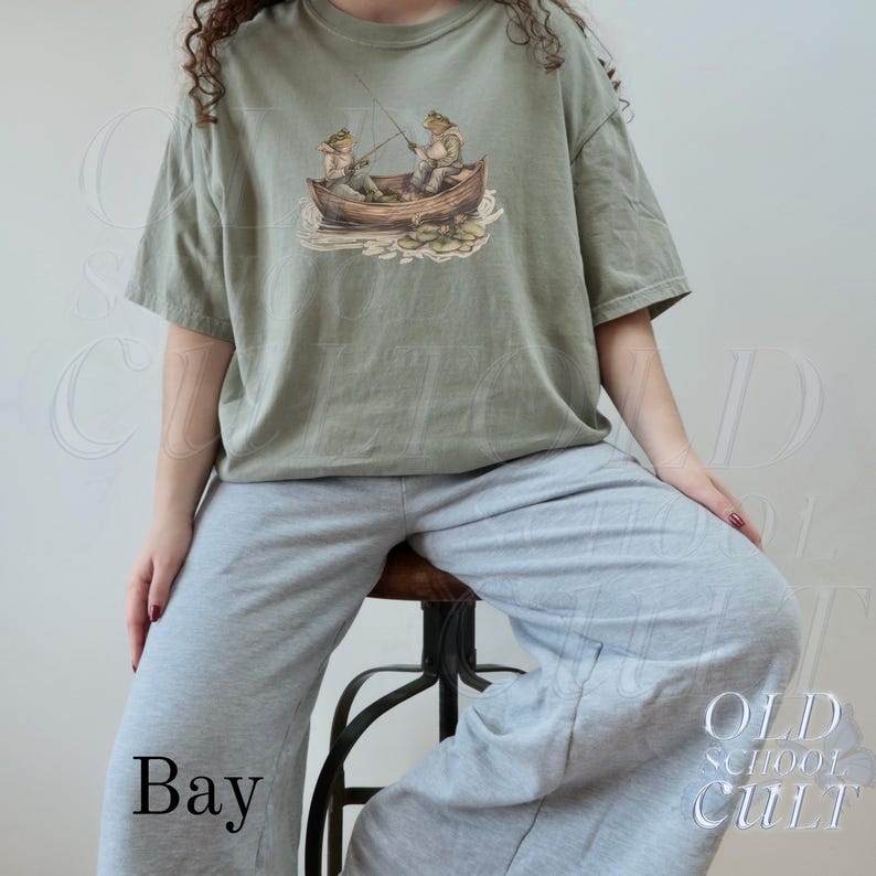 Pode incluir: Camiseta verde s&aacute;lvia com uma ilustra&ccedil;&atilde;o fantasiosa de dois sapos pescando em um barco de madeira. O gr&aacute;fico inclui nen&uacute;fares e varas de pesca. A camiseta &eacute; combinada com cal&ccedil;as de moletom cinza. A palavra "Bay" est&aacute; impressa na parte inferior.