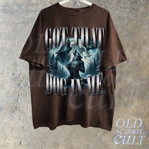 May include: Brown t-shirt with a graphic print of two wolves howling at the moon. The text "GOT THAT DOG IN ME" is printed in silver lettering on the front of the shirt.