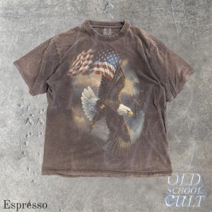 May include: Espresso-coloured t-shirt featuring a bald eagle with an American flag. The eagle is in mid-flight, holding the flag. The shirt has a vintage, distressed look. The text "OLD SCHOOL CULT" is printed on the bottom right.