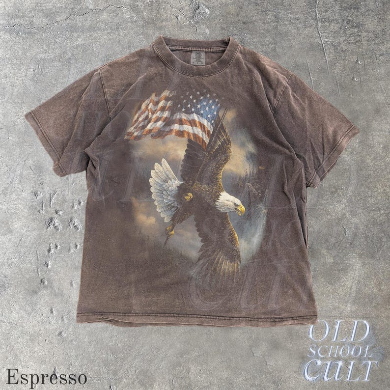May include: Espresso-coloured t-shirt featuring a bald eagle with an American flag. The eagle is in mid-flight, holding the flag. The shirt has a vintage, distressed look. The text "OLD SCHOOL CULT" is printed on the bottom right.