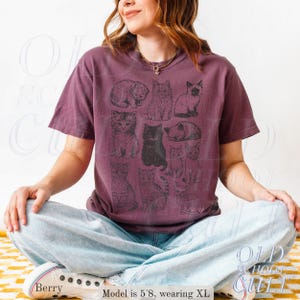 May include: Berry-colored t-shirt featuring a collection of illustrated cats in various poses. The shirt has a vintage, worn look. The model is wearing an XL size.