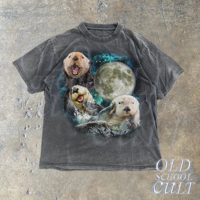 May include: A black t-shirt with a faded graphic of three sea otters swimming in front of a full moon. The otters are smiling and appear to be in a playful mood. The background is a dark blue with stars.
