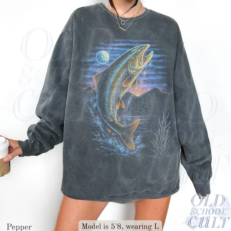 May include: Dark grey sweatshirt with a leaping fish design, moon, and mountain backdrop. The fish is in blue, gold, and brown. The sweatshirt has a vintage, washed look and long sleeves. The text "OLD SCHOOL CULT" is visible.