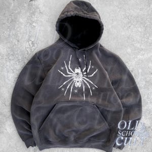 May include: A dark gray hoodie with a large white spider graphic on the front. The hoodie has a drawstring hood and a kangaroo pocket. The words "OLD SCHOOL CULT" are printed on the lower right corner.