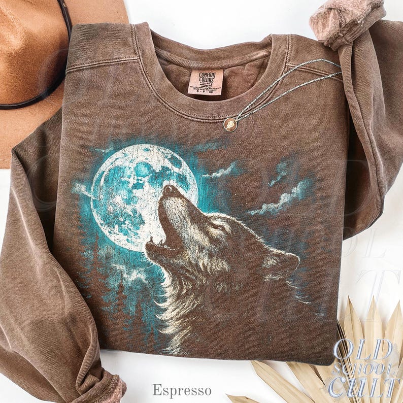 May include: Espresso-colored sweatshirt featuring a howling wolf against a teal-colored moon. The graphic design includes a forest silhouette and clouds. The sweatshirt has a crew neck and long sleeves. A silver necklace with a pendant is draped on the sweatshirt.