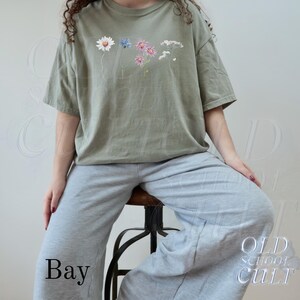 Pu&ograve; includere: T-shirt verde salvia con un design floreale con una margherita, un fiore blu e fiori rosa. La maglietta &egrave; abbinata a pantaloni della tuta grigi. La parola "Bay" &egrave; stampata in basso a sinistra e "OLD SCHOOL CULT" in basso a destra.