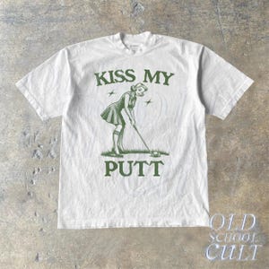 May include: White t-shirt with green text that reads "Kiss My Putt". The graphic features a cartoon woman in a golf outfit putting a golf ball.