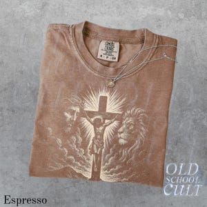 May include: Espresso-colored t-shirt with a graphic of Jesus on the cross, a lion, and a man's face. The shirt has a crew neck and short sleeves. A silver necklace with a pendant rests on the shirt. The text "OLD SCHOOL CULT" is visible.