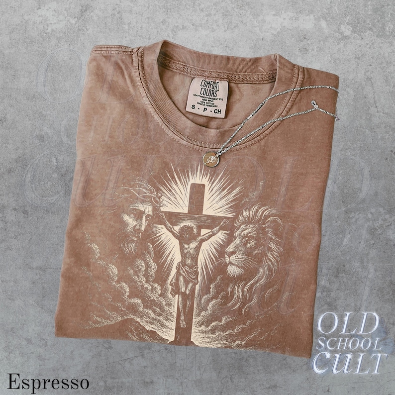 May include: Espresso-colored t-shirt with a graphic of Jesus on the cross, a lion, and a man's face. The shirt has a crew neck and short sleeves. A silver necklace with a pendant rests on the shirt. The text "OLD SCHOOL CULT" is visible.