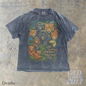 May include: A denim-colored t-shirt featuring a vibrant graphic of various jungle animals, including tigers, monkeys, and a gorilla, surrounded by lush green foliage. The shirt has the text "Life is precious! Take care of it."