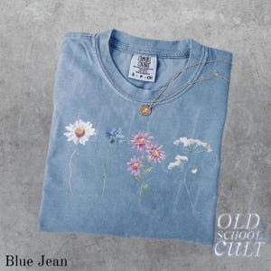 Pu&ograve; includere: Una maglietta blu jeans con un design floreale e una collana d'oro. La maglietta presenta una margherita, un fiore blu, fiori rosa e fiori bianchi. La maglietta ha il testo "OLD SCHOOL CULT" e "Blue Jean".