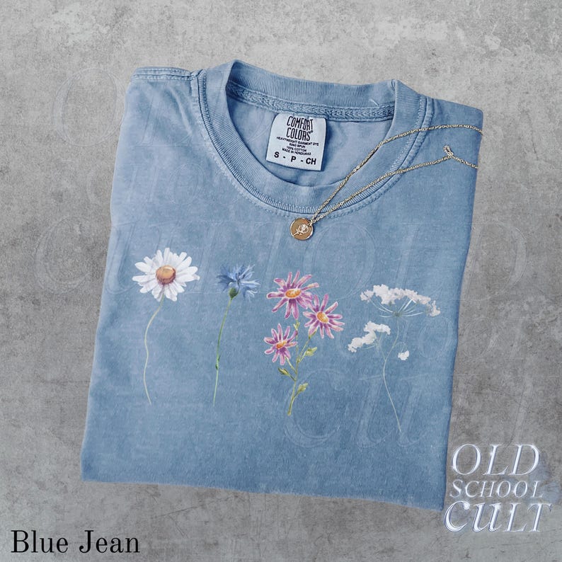 Pu&ograve; includere: Una maglietta blu jeans con un design floreale e una collana d'oro. La maglietta presenta una margherita, un fiore blu, fiori rosa e fiori bianchi. La maglietta ha il testo "OLD SCHOOL CULT" e "Blue Jean".