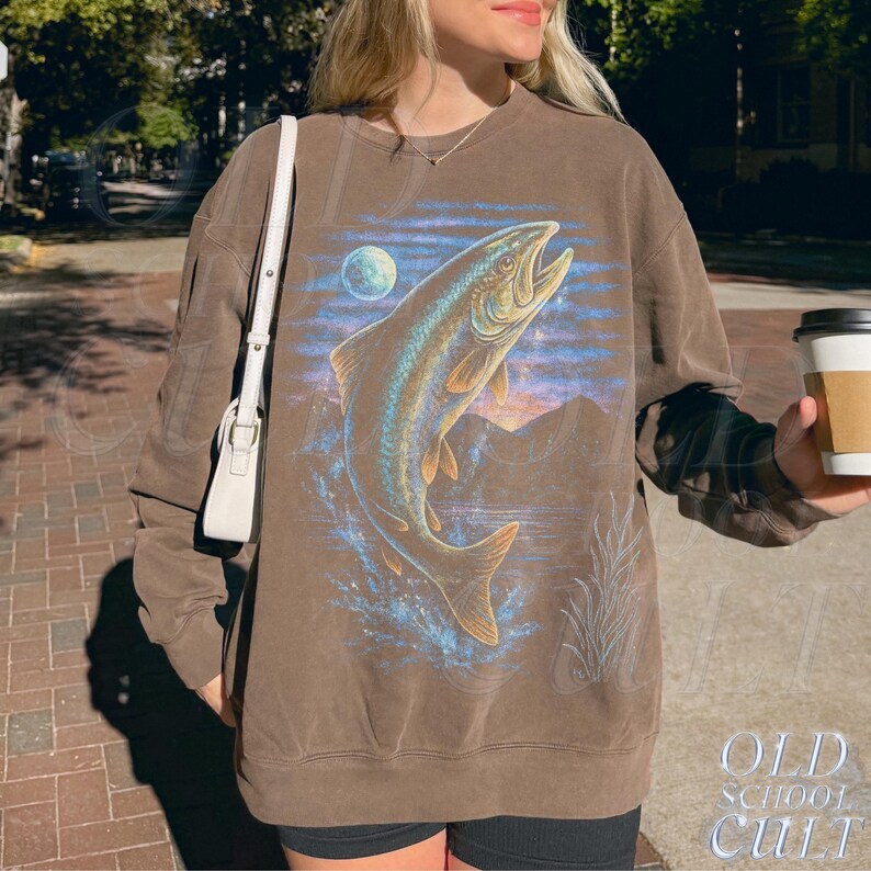 May include: Brown sweatshirt with a leaping fish design, moon, and mountain backdrop. The graphic is in blue, gold, and brown. The text "OLD SCHOOL CULT" is printed in white on the lower right side of the sweatshirt.