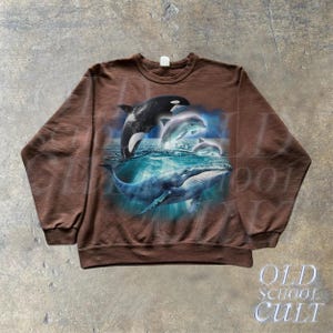 May include: A brown sweatshirt with a graphic print of a whale and dolphins jumping out of the water. The graphic is in shades of blue and white.