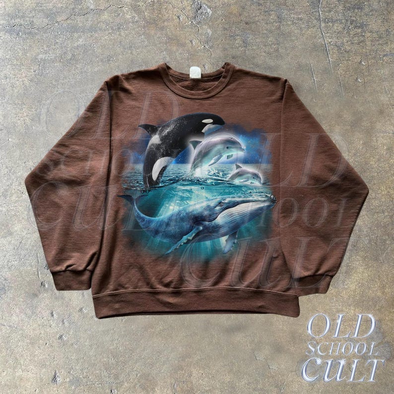 May include: A brown sweatshirt with a graphic print of a whale and dolphins jumping out of the water. The graphic is in shades of blue and white.