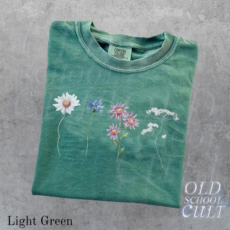 Pu&ograve; includere: T-shirt verde chiaro con un design floreale con una margherita bianca, un fiordaliso blu, margherite rosa e viola e fiori di campo bianchi. La maglietta ha il testo "OLD SCHOOL CULT" e "Light Green".