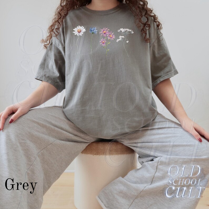 Pu&ograve; includere: Una maglietta grigia con un design floreale con fiori bianchi, blu e rosa. La maglietta &egrave; abbinata a pantaloni della tuta grigi. Il testo "Grey" si trova in basso a sinistra nell'immagine e "OLD SCHOOL CULT" in basso a destra.