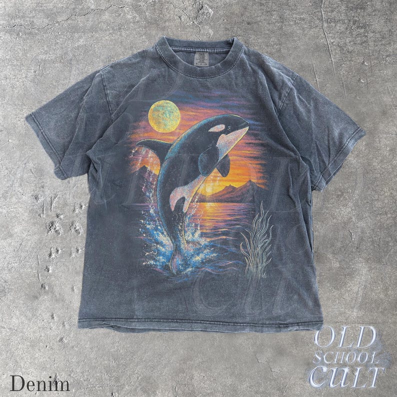 Puede incluir: Camiseta azul denim con un gr&aacute;fico de una orca saltando sobre un fondo de puesta de sol y luna llena. La camiseta tiene un aspecto vintage y descolorido. El texto "OLD SCHOOL CULT" est&aacute; impreso en la parte inferior derecha.