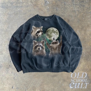 Op de afbeelding: Een zwart sweatshirt met een grafische print van drie wasberen die naar een volle maan reiken. De wasberen bevinden zich in een bosrijke omgeving met groen gebladerte.