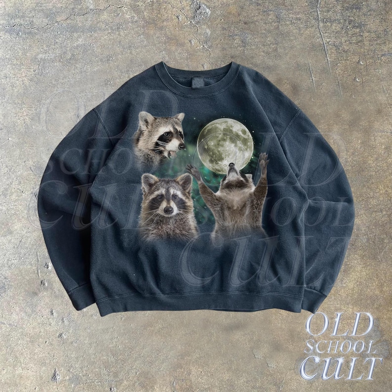 Op de afbeelding: Een zwart sweatshirt met een grafische print van drie wasberen die naar een volle maan reiken. De wasberen bevinden zich in een bosrijke omgeving met groen gebladerte.
