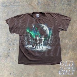 May include: A brown t-shirt with a graphic print of two wolves howling at the moon. The wolves are surrounded by a green and blue aurora borealis. The shirt has a faded look and is made of a soft, comfortable material.
