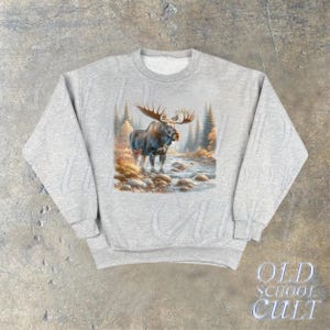 May include: A gray sweatshirt with a graphic print of a moose standing in a forest setting. The moose is brown and has large antlers. The forest is green and brown with a stream running through it.