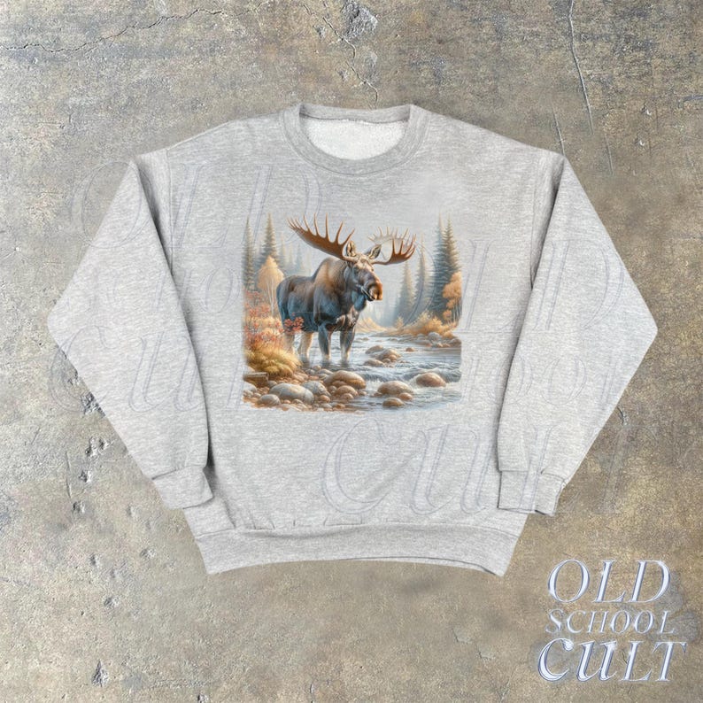 May include: A gray sweatshirt with a graphic print of a moose standing in a forest setting. The moose is brown and has large antlers. The forest is green and brown with a stream running through it.