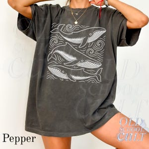 May include: A dark gray t-shirt featuring a white graphic of three whales within a square frame, surrounded by wave-like designs. The word "Pepper" is printed in the lower left corner. The shirt has short sleeves and a relaxed fit.