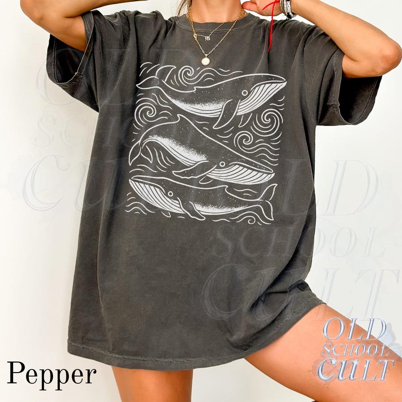 May include: A dark gray t-shirt featuring a white graphic of three whales within a square frame, surrounded by wave-like designs. The word "Pepper" is printed in the lower left corner. The shirt has short sleeves and a relaxed fit.