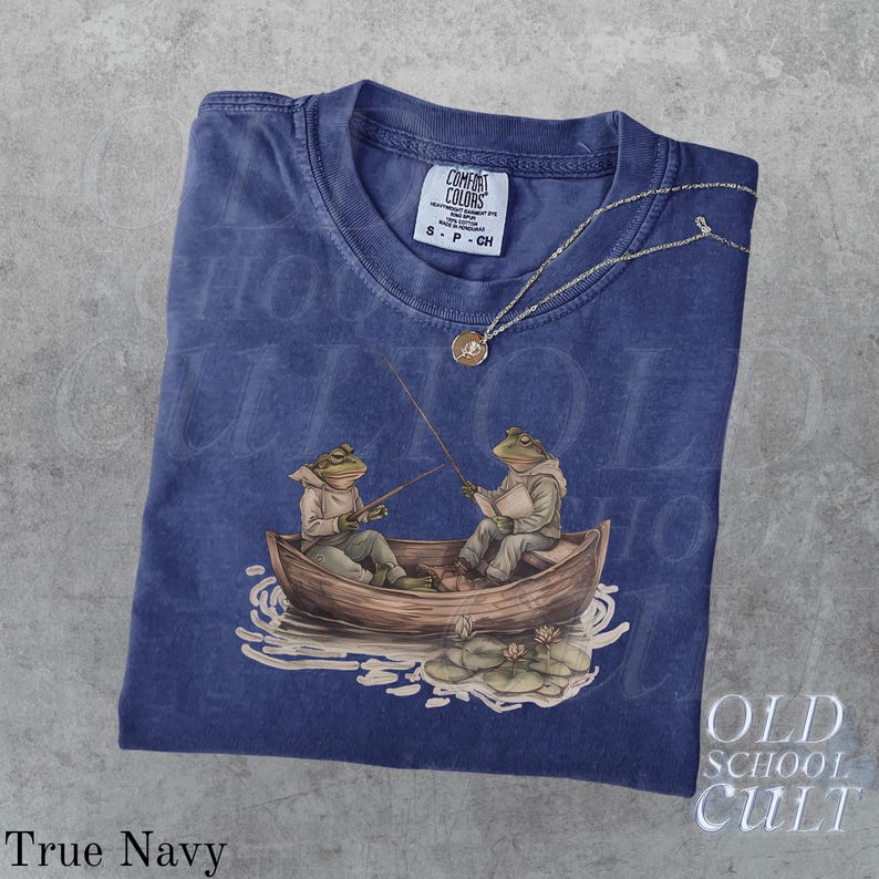 Pode incluir: Camiseta azul marinho com um gr&aacute;fico de dois sapos pescando em um barco. A camiseta tem gola redonda e uma pequena etiqueta na gola. Um colar de prata com um pingente est&aacute; drapeado sobre a camiseta. As palavras "Old School Cult" s&atilde;o impressas no canto inferior direito.