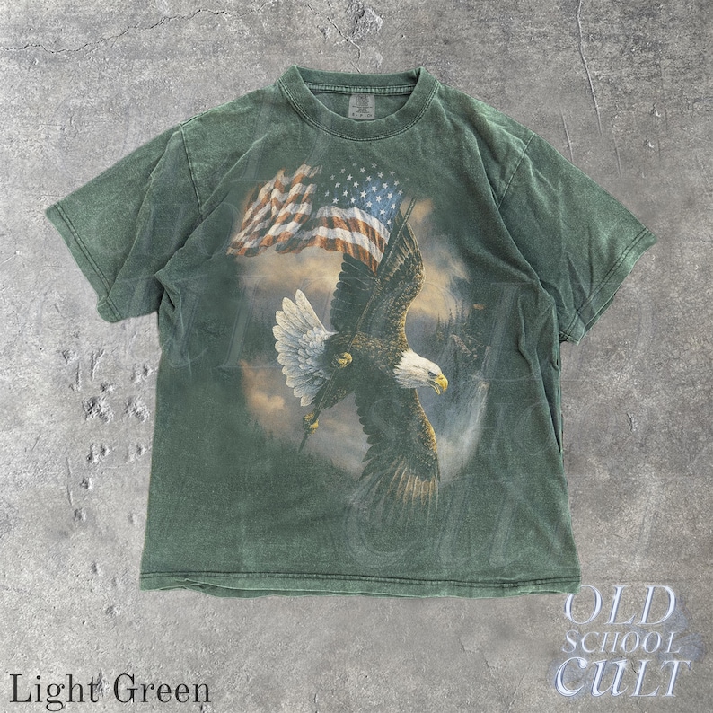May include: Light green t-shirt with a graphic of an eagle and the American flag. The eagle is in flight. The shirt has a faded appearance. The text "OLD SCHOOL CULT" is printed on the bottom right. The words "Light Green" are printed on the bottom left.