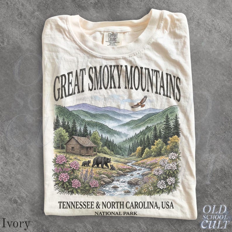 K&ouml;nnte beinhalten: Ein elfenbeinfarbenes T-Shirt mit einem Grafikdruck der Great Smoky Mountains. Das Motiv zeigt eine Blockh&uuml;tte, einen Bach, Schwarzb&auml;ren und einen fliegenden Adler inmitten von Kiefern und Blumen, mit dem Text Great Smoky Mountains und Tennessee & North Carolina, USA National Park.
