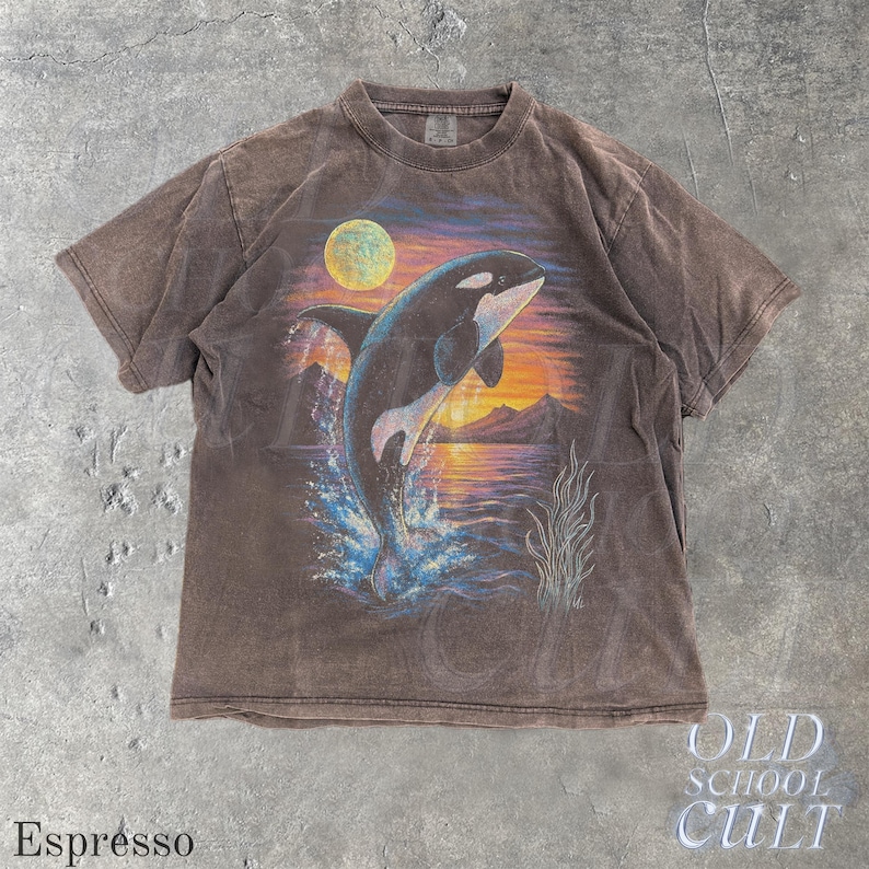 Puede incluir: Camiseta color espresso con un gr&aacute;fico de una orca saltando sobre un fondo de puesta de sol. El dise&ntilde;o incluye una luna llena, monta&ntilde;as y agua. La camiseta tiene un aspecto vintage y desgastado, con el texto "OLD SCHOOL CULT" en la esquina inferior derecha.