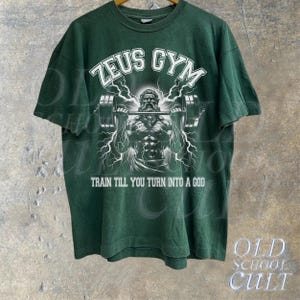 May include: Green t-shirt with a white graphic of a muscular figure lifting a barbell. The graphic includes the text "Zeus Gym" and "Train till you turn into a god."