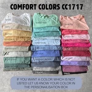 May include: A display of Comfort Colors CC1717 t-shirts in various colors, including white, ivory, and peachy. The image features stacks of folded shirts with color names like "island reef" and "berry" displayed. Text at the top reads "COMFORT COLORS CC1717".