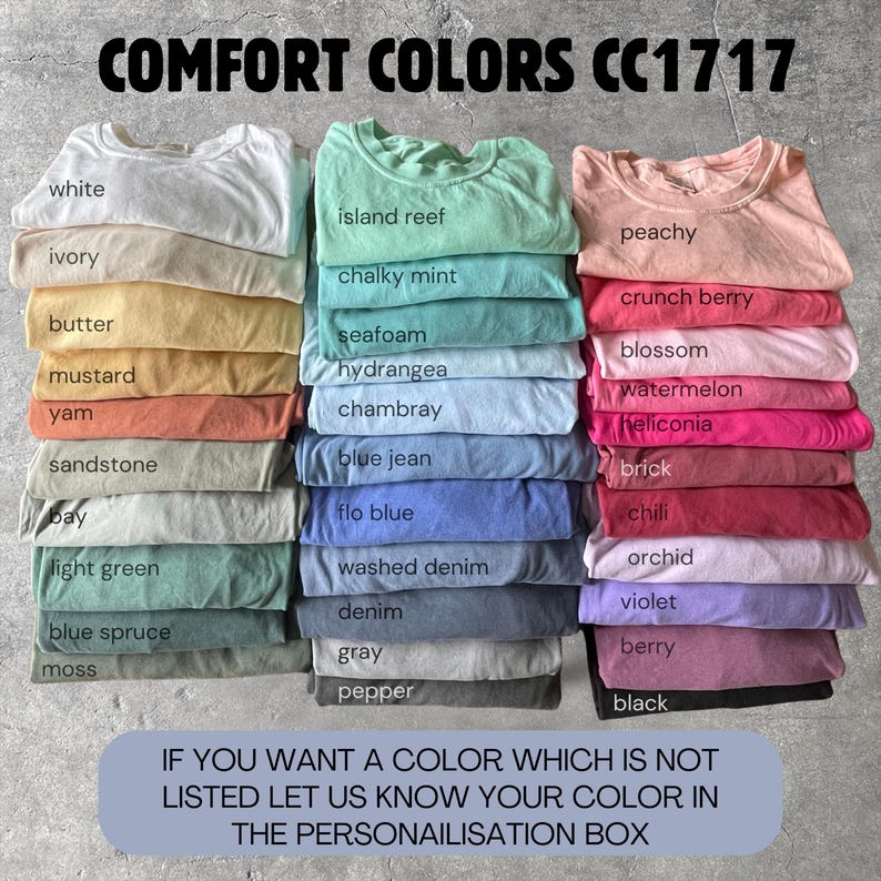 May include: A display of Comfort Colors CC1717 t-shirts in various colors, including white, ivory, and peachy. The image features stacks of folded shirts with color names like "island reef" and "berry" displayed. Text at the top reads "COMFORT COLORS CC1717".